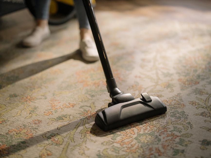 Close-up view of a vacuum cleaner head actively cleaning a patterned, plush carpet with floral designs in warm tones. The vacuum head is black with a small switch on top, connected to a long, black ex