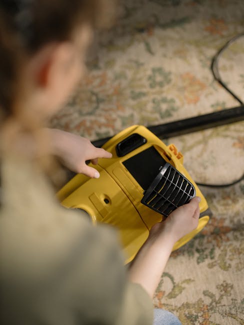 A woman is performing surface cleaning on a patterned area rug with a handheld vacuum cleaner in a living room. The rug features intricate floral designs in muted shades of beige, green, and orange, s